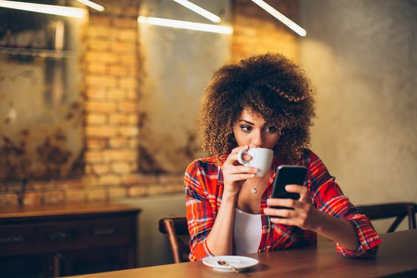 زن جوان در کافی نت که قهوه می نوشد و از تلفن همراه استفاده می کند