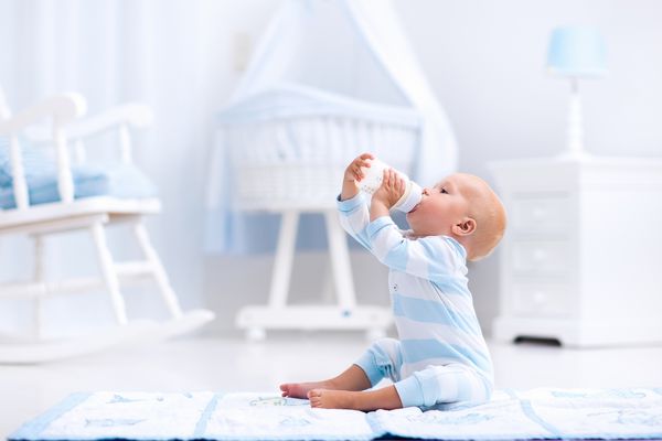 پسر بچه شایان ستایش بر روی یک تشک کف آبی و نوشیدن شیر از بطری در یک مهد کودک سفید آفتابی با صندلی تکان و بیس بال فضای داخلی اتاق خواب با گهواره نوزاد نوشیدنی فرمول برای نوزاد