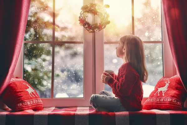 کریسمس مبارک و تعطیلات مبارک دختر بچه ناز که کنار پنجره با یک لیوان نوشیدنی نشسته و به جنگل زمستانی نگاه می کند اتاق تزئین شده در کریسمس بچه از بارش برف لذت می برد