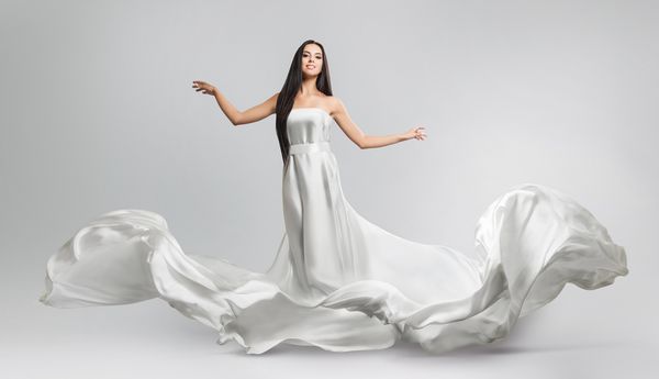 دختر جوان زیبا در لباس سفید پرواز پارچه در حال جریان پارچه سفید سبک که در باد پرواز می کند