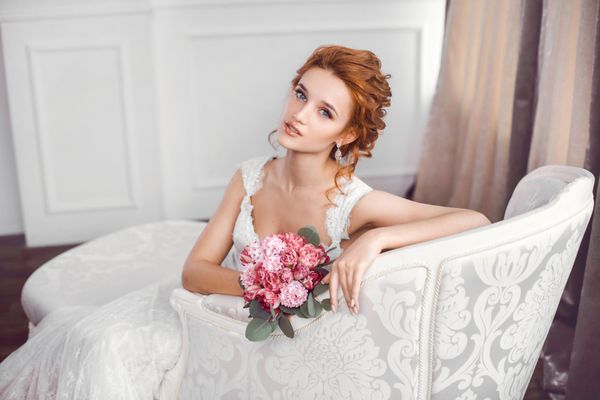 عروس با لباس زیبا که روی مبل در داخل خانه با دسته گلهایی که در فضای استودیوی سفید مانند در خانه است نشسته است شیک عروسی مرسوم مد روز به طول کامل مدل موهای جذاب قرمز قفقازی جوان دوست دارند