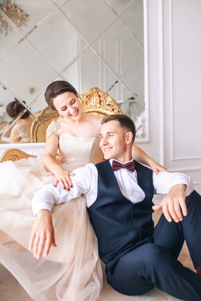 عروس با لباس زیبا و داماد با کت و شلوار مشکی که بر روی کاناپه در فضای داخلی استودیوی سفید مانند در خانه نشسته است لباس عروس مرسوم مد روز داماد در آغوش گرفتن عروس