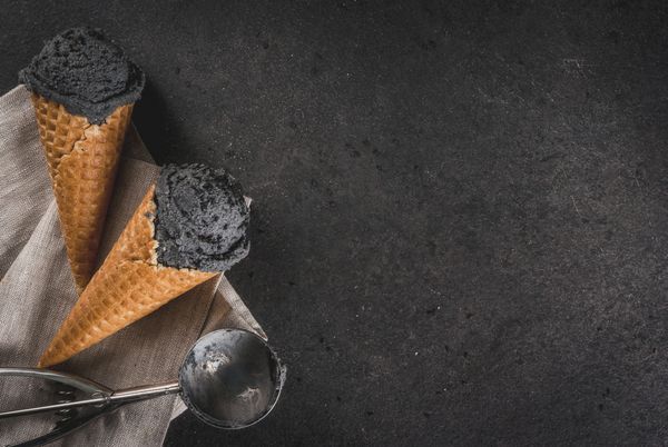 غذای مرسوم مد روز بستنی مشکی با کنجد سیاه در مخروط های بستنی سنتی به شکل مخلوط شده روی یک میز سنگی سیاه در یک سینی چوبی نمای نمای فضا را کپی کنید