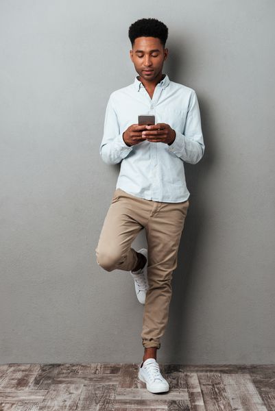 پرتره طول کامل یک مرد جوان آمریکایی آفریقایی در حال تایپ پیام در تلفن همراه در حالی که در حال نمایش و ایستادن بر روی زمینه خاکستری است