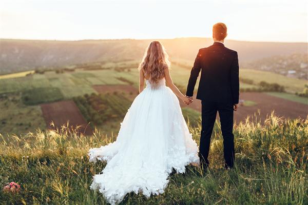 عروسی عروس و داماد دست در دست هم ایستاده اند که در دامنه تپه ایستاده اند و به غروب خورشید نگاه می کنند