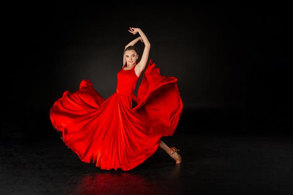 عکس استودیویی از دختر زیبای اروپایی با لباس قرمز در حال رقص با دستان بلند شده در استودیو در پس زمینه خاکستری