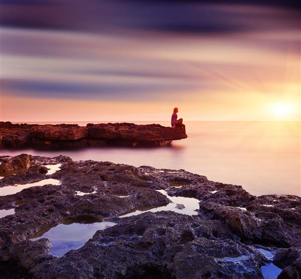 دختری که روی صخره ای کنار دریا نشسته و غروب زیبای آفتاب را تماشا می کند