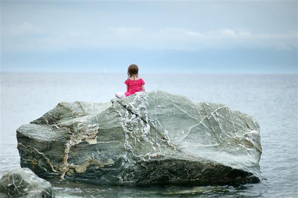 دختر کوچک که روی صخره ای کنار دریا نشسته است