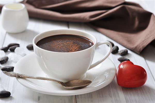 شکلات داغ در فنجان با پودر کاکائو و قلب سنگ قرمز در زمینه چوبی