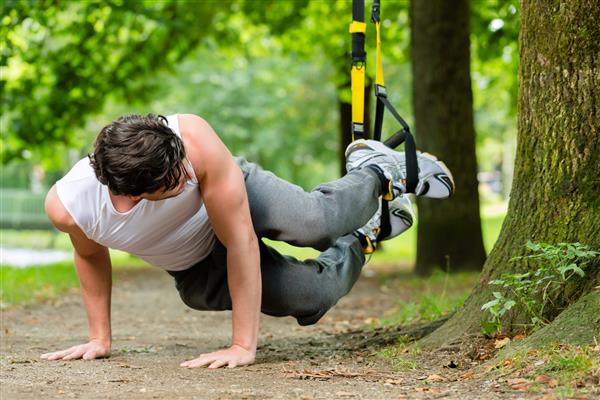 مرد جوانی در حال ورزش با زنجیر مربی سیستم تعلیق در شهر پارک زیر درختان تابستان برای تناسب اندام ورزشی است