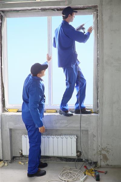 دو مرد با لباس کار قاب پنجره جدیدی تنظیم می کنند یکی از آنها روی طاقچه ایستاده و قاب را محکم می کند