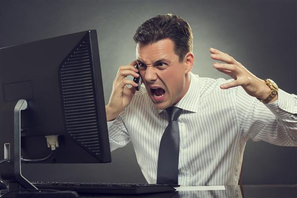 یک تاجر عصبانی با پیراهن و گردن سفید در حین مکالمه تلفن همراه در مقابل صفحه نمایش رایانه و در پس زمینه خاکستری فریاد و اشاره می کند