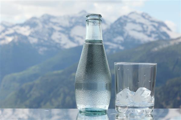بطری آب معدنی و لیوان با مکعب های یخ کوه های آلپ در پس زمینه