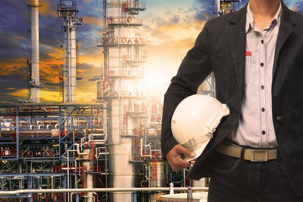 مرد مهندسی با کلاه ایمنی سفید در مقابل ساختمان ساختمان پالایشگاه نفت در صنایع سنگین پتروشیمی ایستاده است
