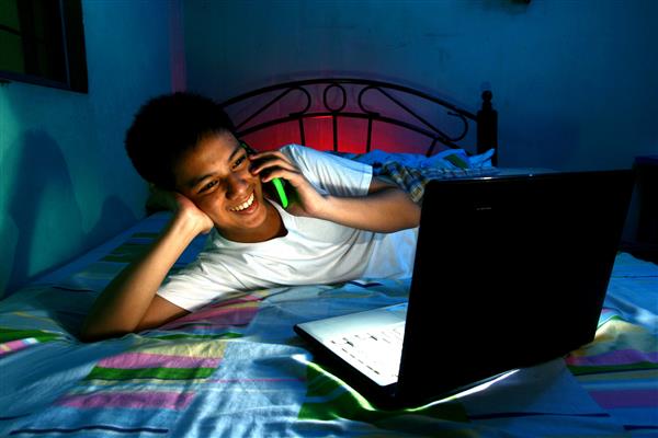 نوجوان جوان جلوی رایانه لپ تاپ و روی تخت و استفاده از تلفن همراه یا تلفن هوشمند عکس نوجوان جوان مقابل رایانه لپ تاپ و روی تخت و استفاده از تلفن همراه یا تلفن هوشمند