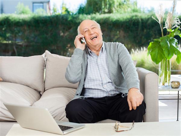 یک مرد ارشد شاد و خنده در حالی که در حال پاسخ به تلفن هوشمند روی کاناپه در ایوان خانه سالمندان است