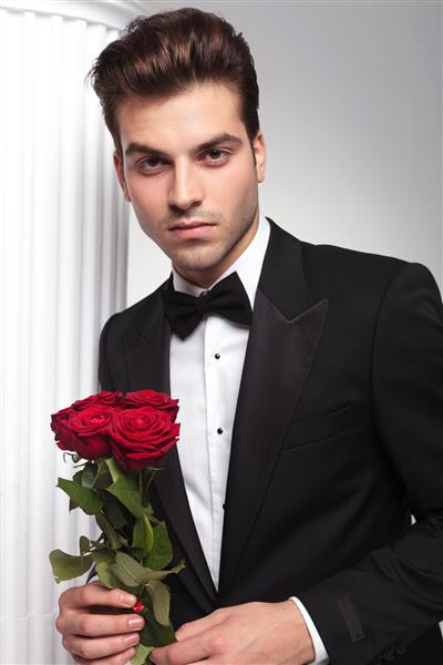 مرد خوش تیپ و تکیه به ستونی سفید در حالی که گل رزهای قرمز را در دست گرفته و به دوربین نگاه می کند
