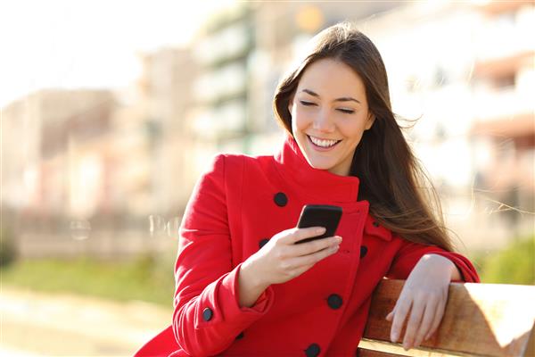 دختری که با تلفن هوشمند پیامکی می کند که در پارک نشسته و ژاکت قرمز بر تن دارد و در یک نیمکت در پارک نشسته است