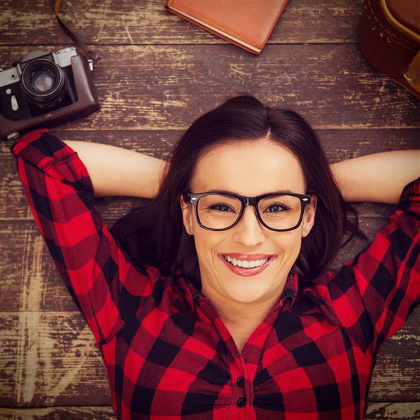 رویای یک سفر بزرگ نمای بالایی از زن جوان زیبا با عینکی که روی زمین چوب سخت قرار گرفته و در حالی که چمدان و دوربین در نزدیکی او خوابیده است لبخند می زند