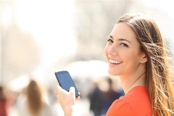 زن خوشبختی که با استفاده از تلفن هوشمند در خیابان لبخند می زند و در خیابان قدم می زند و به دوربین نگاه می کند