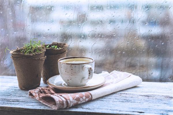 فنجان با نوشیدنی گرم روی میز چوبی جلوی پنجره با قطرات باران هوای بارانی طبیعت بی روح تن های کمرنگ و سرد تصویر افقی