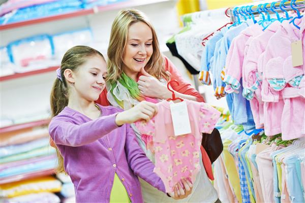 زن باردار و دختر کوچک هنگام خرید از سوپرمارکت پوشاک لباس نوزاد تازه متولد شده را انتخاب می کنند