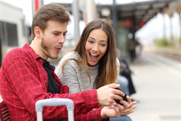 زن و شوهر خنده دار در حالی که منتظر هستند با یک تلفن هوشمند در ایستگاه قطار بازی می کنند