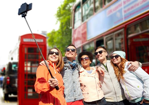 جهانگردی مسافرت مردم اوقات فراغت و مفهوم فناوری - گروهی از دوستان نوجوان خندان که با تلفن های هوشمند و مونوپاد از پس زمینه خیابان لندن سلفی می گیرند