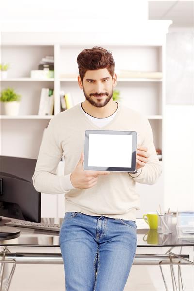 پرتره یک تاجر جوان که همزمان با ایستادن در دفتر و لبخند زدن تبلت دیجیتال را با صفحه سفید در دست گرفته است