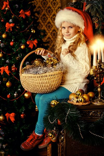 دختر کوچک خوشحال در کلاه سانتا در اتاقی که برای کریسمس تزئین شده نشسته است