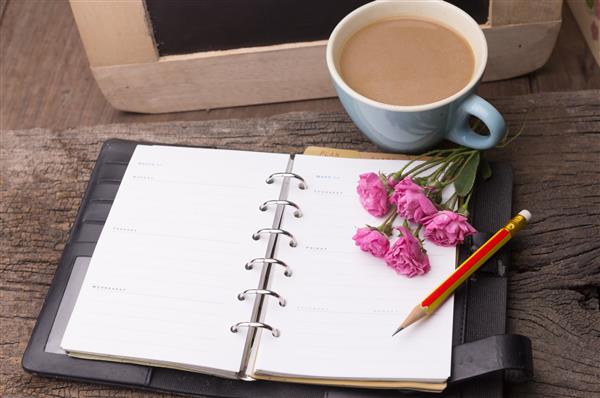 گل رز صورتی لیوان با قهوه دفتر خاطرات و مداد روی میز چوبی