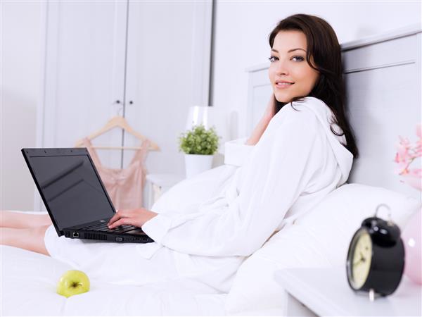 زن زیبا و بزرگسال جوان در اتاق خواب با لپ تاپ روی تخت نشسته است