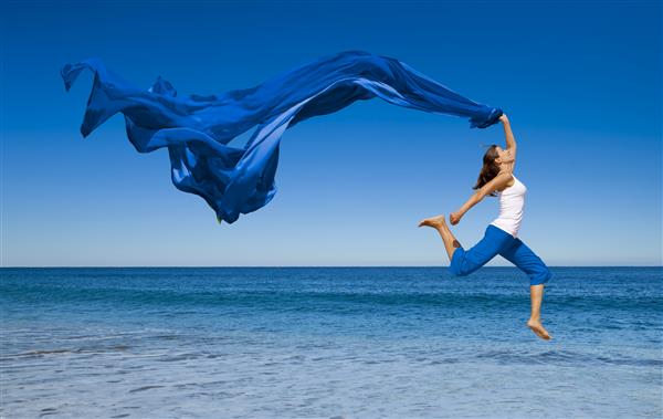 زن جوان زیبا با دستمال رنگی در حال پریدن در ساحل است
