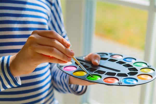 بچه کوچولو در مدرسه با نقاشی آبرنگ نقاشی می کند قلم موی آغشته را به رنگ رنگی نزدیک می کند
