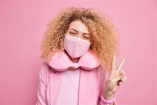 زن جوان بی دغدغه و خوشحال از ماسک برای محافظت در برابر ویروس کرونا استفاده می کند عادت به اندازه گیری قرنطینه باعث می شود علامت صلح با لباس های زیبا از بالش گردن برای احساس راحتی استفاده کند