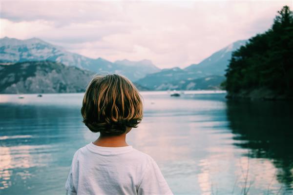بچه ای با موهای بلوند به دریا نگاه می کند با کوه هایی در فاصله دور از پشت عکس می گیرد