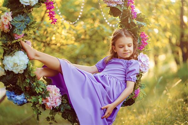 دختر بچه ناز با پوشیدن لباس بنفش در حالیکه در تابستان در حلقه ای تزئین شده با گل نشسته است