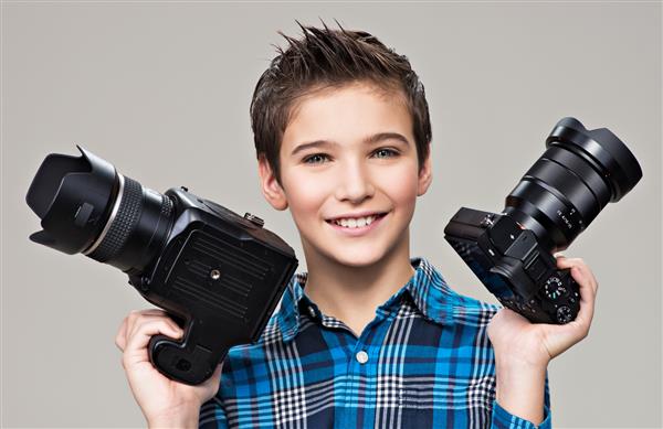 پسر دو دوربین عکاسی را در دست دارد پسر قفقازی خندان با دوربین dslr که در استودیو روی پس‌زمینه خاکستری ژست گرفته است