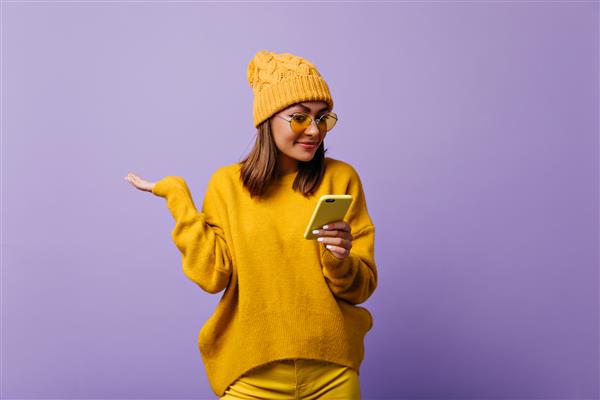 دانش آموز دختر دلپذیر از اروپا با تعجب به پیامک در تلفن خود نگاه می کند دختری با کلاه گرم زرد و عینک های رنگارنگ که برای پرتره ایزوله ژست گرفته است