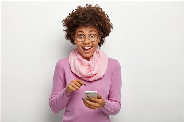 زن با ظاهری دلپذیر با موهای ترد اشاره به دستگاه تلفن همراه دانلود اپلیکیشن مدرن جدید دارای حالتی شاد عینک برای اصلاح بینایی جامپر بنفش و روسری ابریشمی