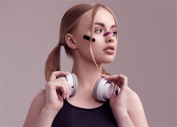 پرتره دختر بلوند زیبا با گچ بری های زرق و برق روی صورتش در حال گوش دادن به موسیقی با هدفون سفید در استودیو