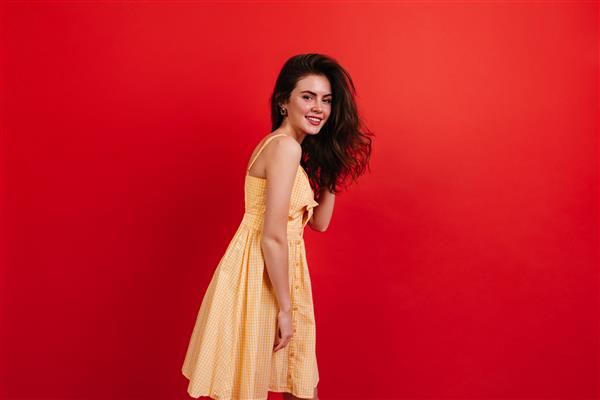 دختر جوان سبزه روی دیوار قرمز ژست گرفته است زن مثبت با لباس زرد زیبا و خندان