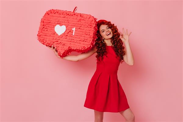 زن شاد با لباس قرمز مینیمالیستی لبخند می زند و علامت ok را نشان می دهد دختری که کلاه بر تن دارد مانند علامت روی فضای صورتی نگه می دارد