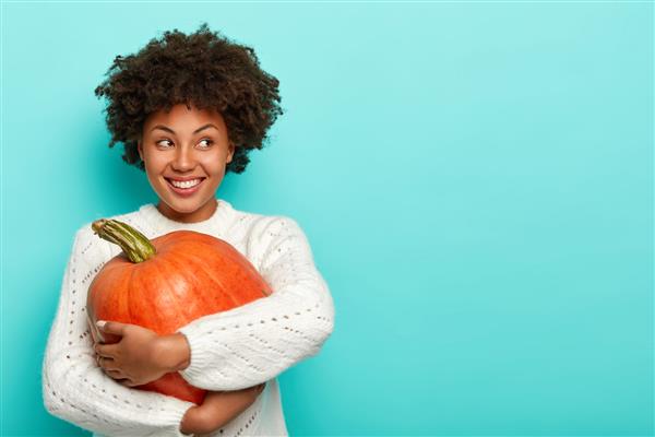 زن خوشحالی که مدل موی آفریقایی دارد کدو تنبل بزرگ در دست دارد از محصول سالم برای تهیه غذای ارگانیک استفاده می کند با خوشحالی به نظر می رسد ژاکت پوشیده است