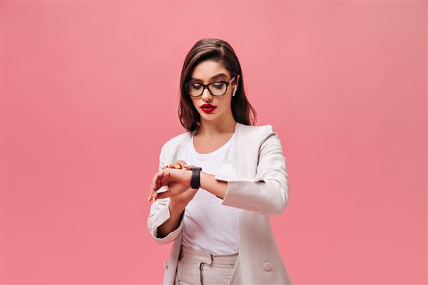 زن شلوغ با ژاکت بژ به ساعت دستی خود نگاه می کند سبزه با لب‌های قرمز روشن با عینک و هدفون سفید که در پس‌زمینه مجزا ژست گرفته است