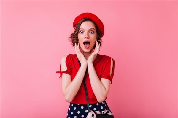 دختر زیبای غافلگیر شده با کلاه فرنگی فرانسوی که با دهان باز ژست گرفته است خانم قفقازی با بلوز قرمز زیبا ایستاده است