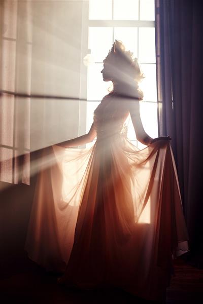 زن بلوند زیبا که زیر نور خورشید پشت پنجره ایستاده است