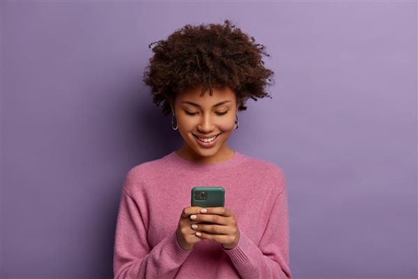 زن زیبای فرفری تلفن همراه مدرن را در دست دارد پیام‌ها را روی دستگاه تلفن هوشمند تایپ می‌کند از ارتباط آنلاین لذت می‌برد برنامه مخصوص چت را دانلود می‌کند با مهربانی لبخند می‌زند روی دیوار بنفش جدا شده است