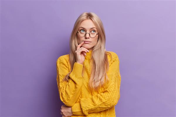 زن جوان و زیبای جوان به ایده های متمرکز در بالا فکر می کند متفکر می ایستد و دست روی صورت می ایستد در حالت متفکرانه می ایستد ژاکت زرد عینک گرد می پوشد