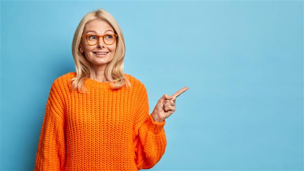 بلوند متفکر زن اروپایی چهل ساله با عینک و ژاکت نارنجی بافتنی که به فضای کپی اشاره می کند